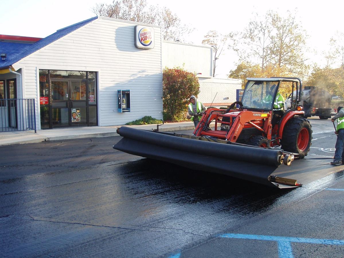 Asphalt fabric overlay installation — a geotextile fabric interlayer being unrolled across a parking lot before the new asphalt overlay is placed