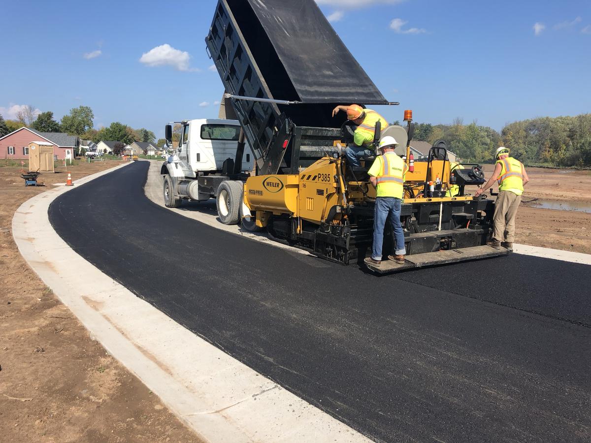 Municipality asphalt paving project — a paving crew and dump truck finishing a new road in a residential subdivision