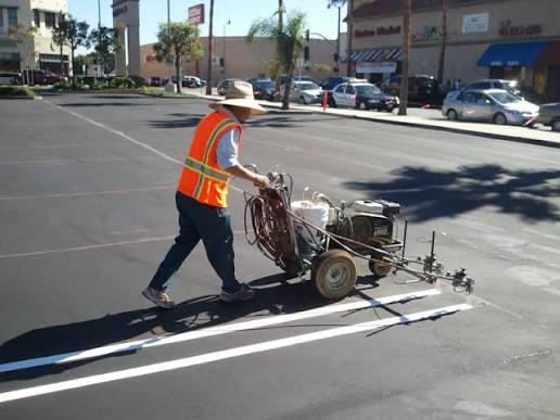 Parking lot maintenance in progress — a worker operating a striping machine to repaint lines in a commercial lot